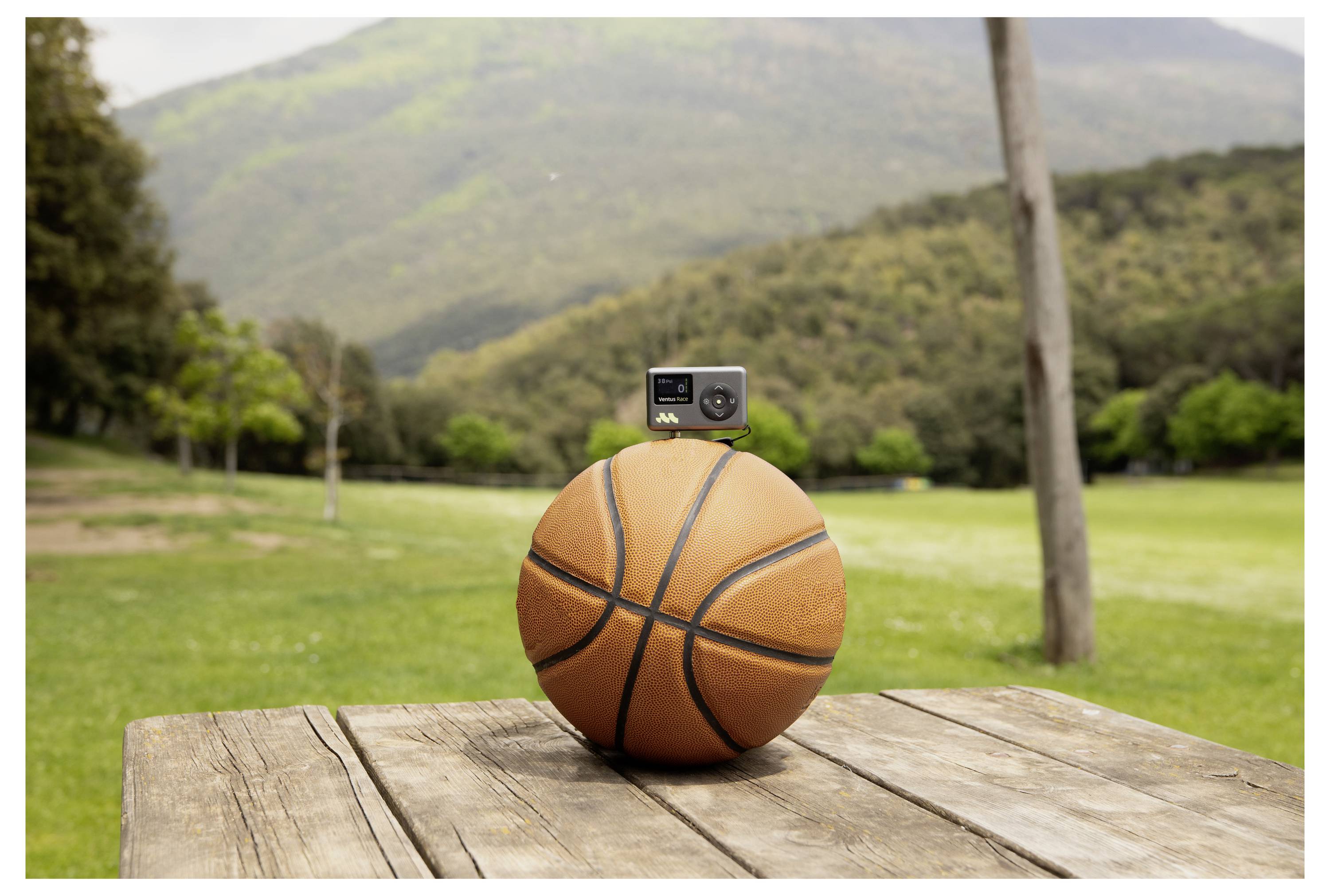 Basketbalová lopta s malou kamerou namontovanou na vrchu, sediaca na drevenom stole vonku s horami a zelenými rastlinami v pozadí.