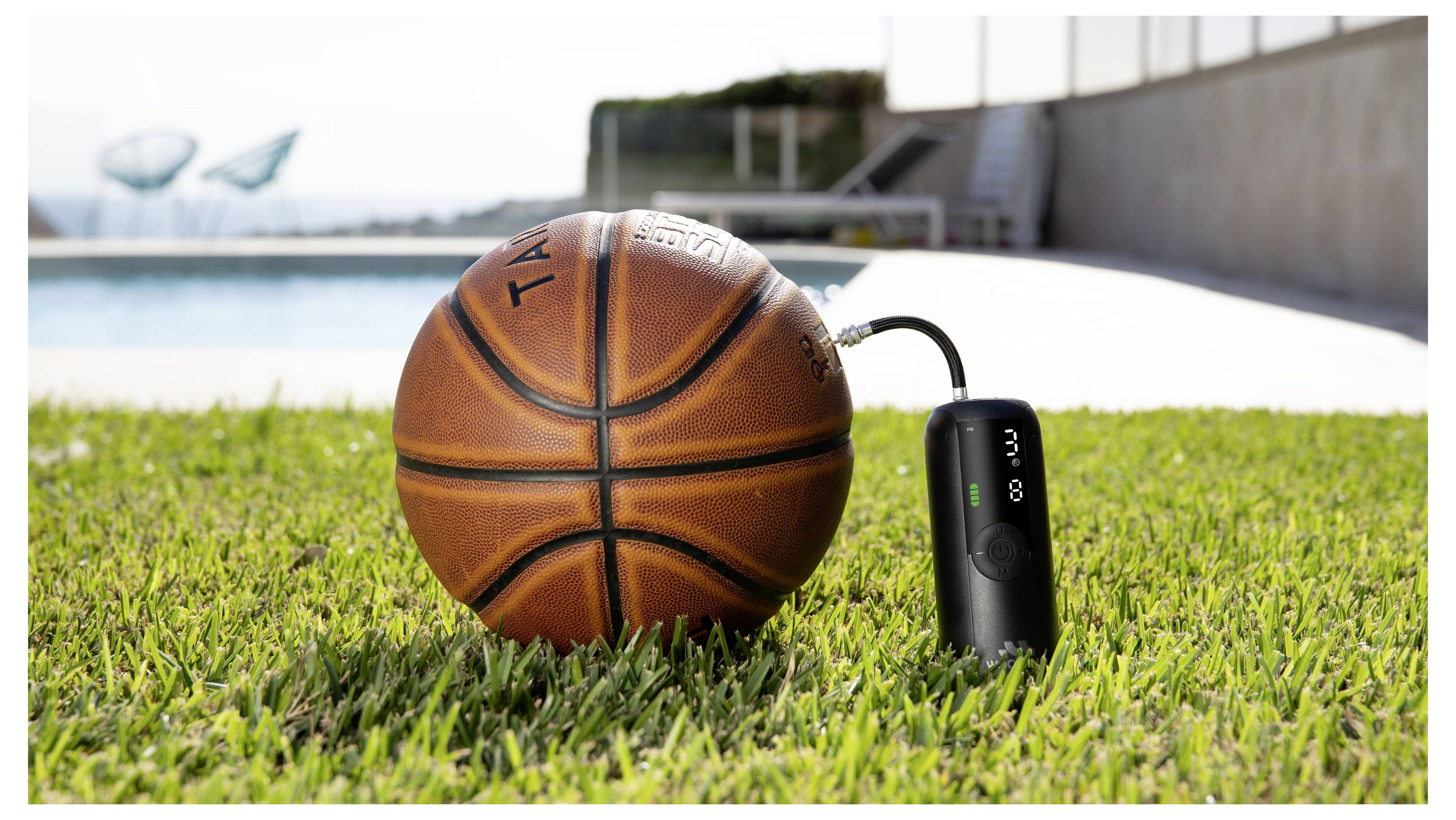 Basketbalová lopta ležiaca na tráve vedľa malého čierneho zariadenia s bazénom a vonkajšou chôdzou v pozadí.
