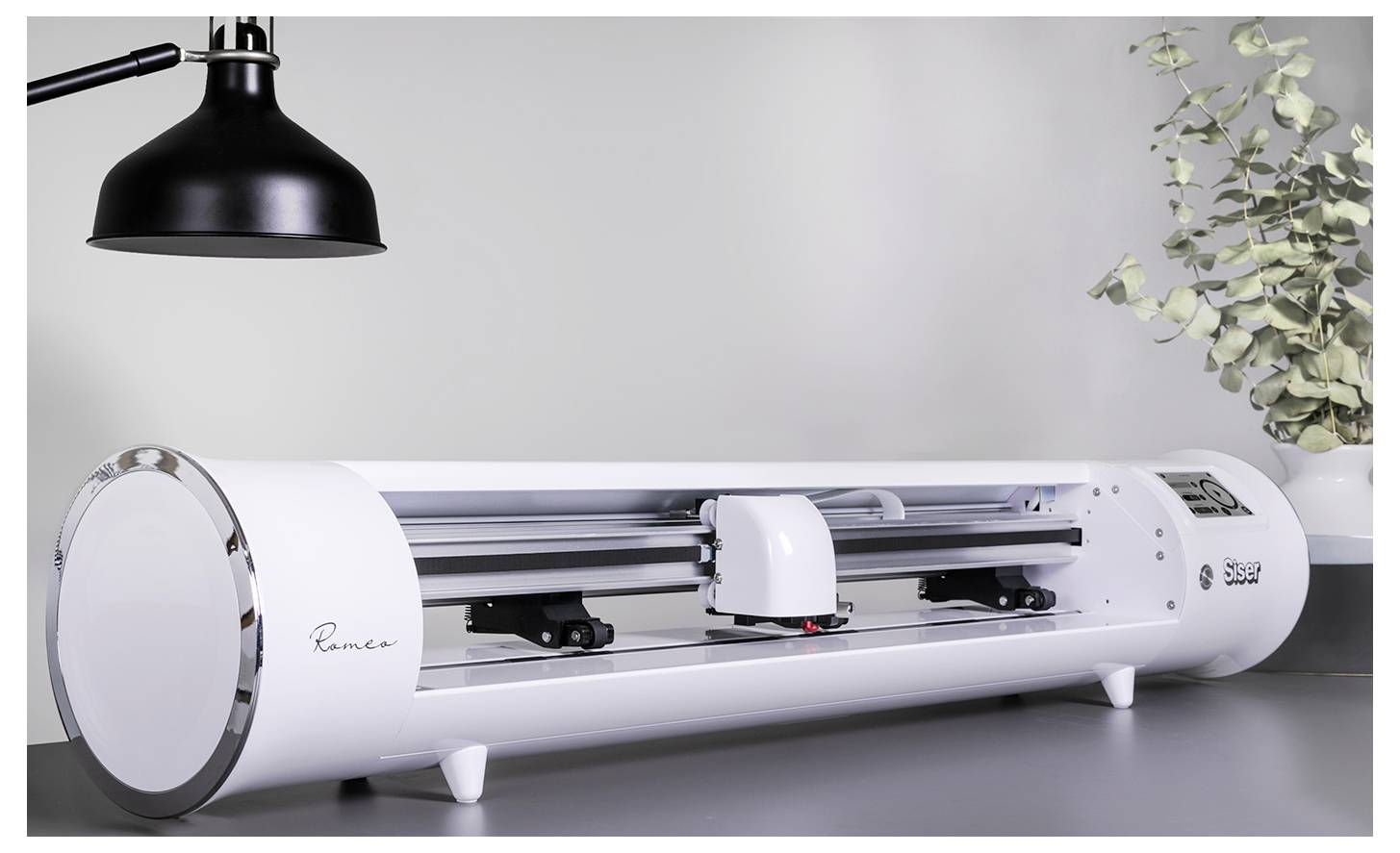 Elegantný, moderný elektronický rezací stroj na stole, s čiernou lampou hore a vetvičkami eukalyptu v blízkosti, v jasnom priestore.