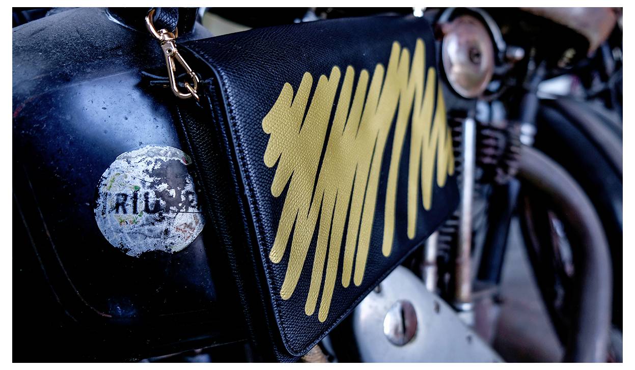 Zblízka fotografia motocykla s čiernym koženým doplnkom s žltými abstraktnými ťahmi štetca, pripevneným na boku motocykla.