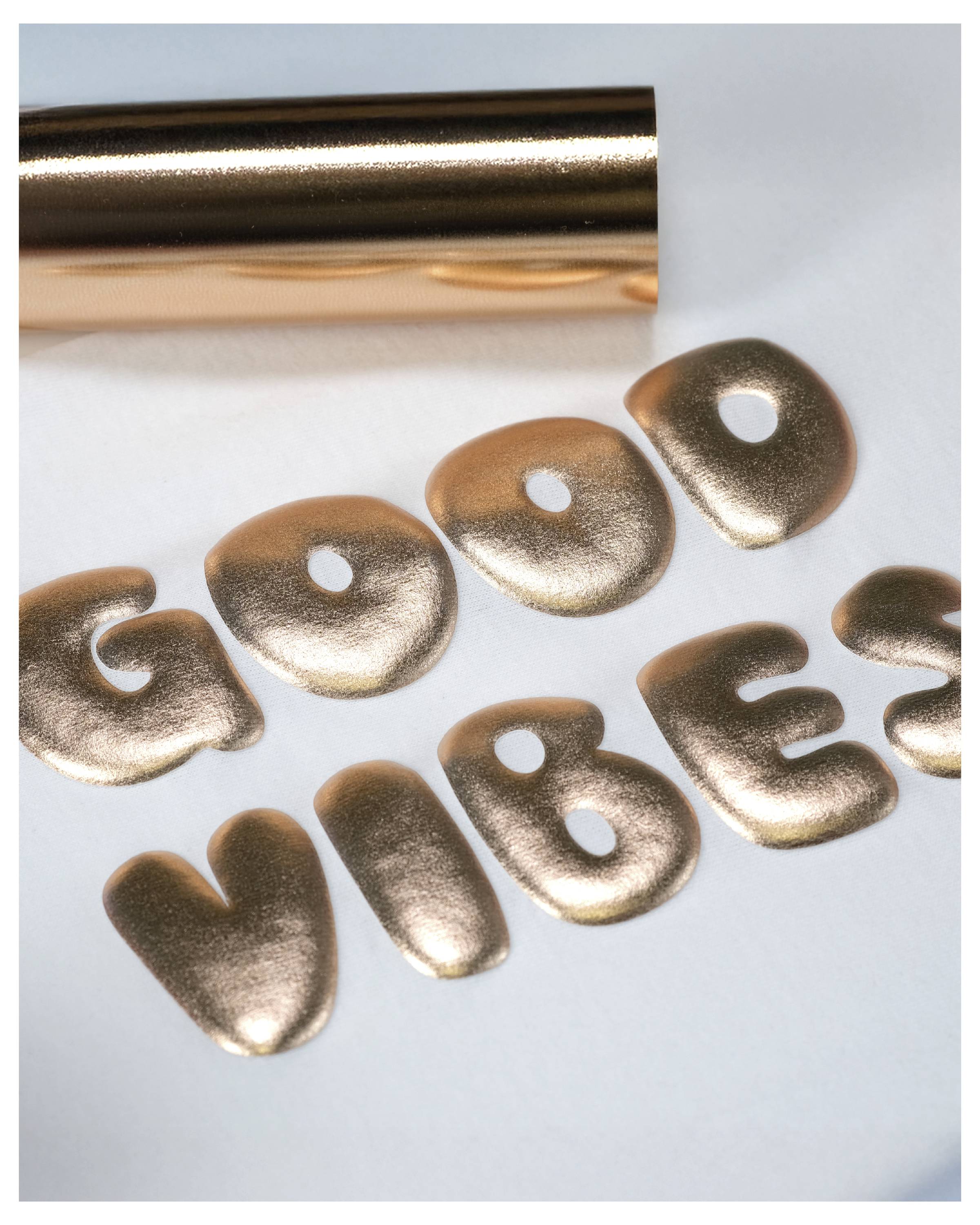 Obrázok zobrazuje zlaté kovové písmená vyskladané do slov "GOOD VIBES" na svetlom pozadí s čiastočne viditeľným rolom zlatej fólie v hornej časti.