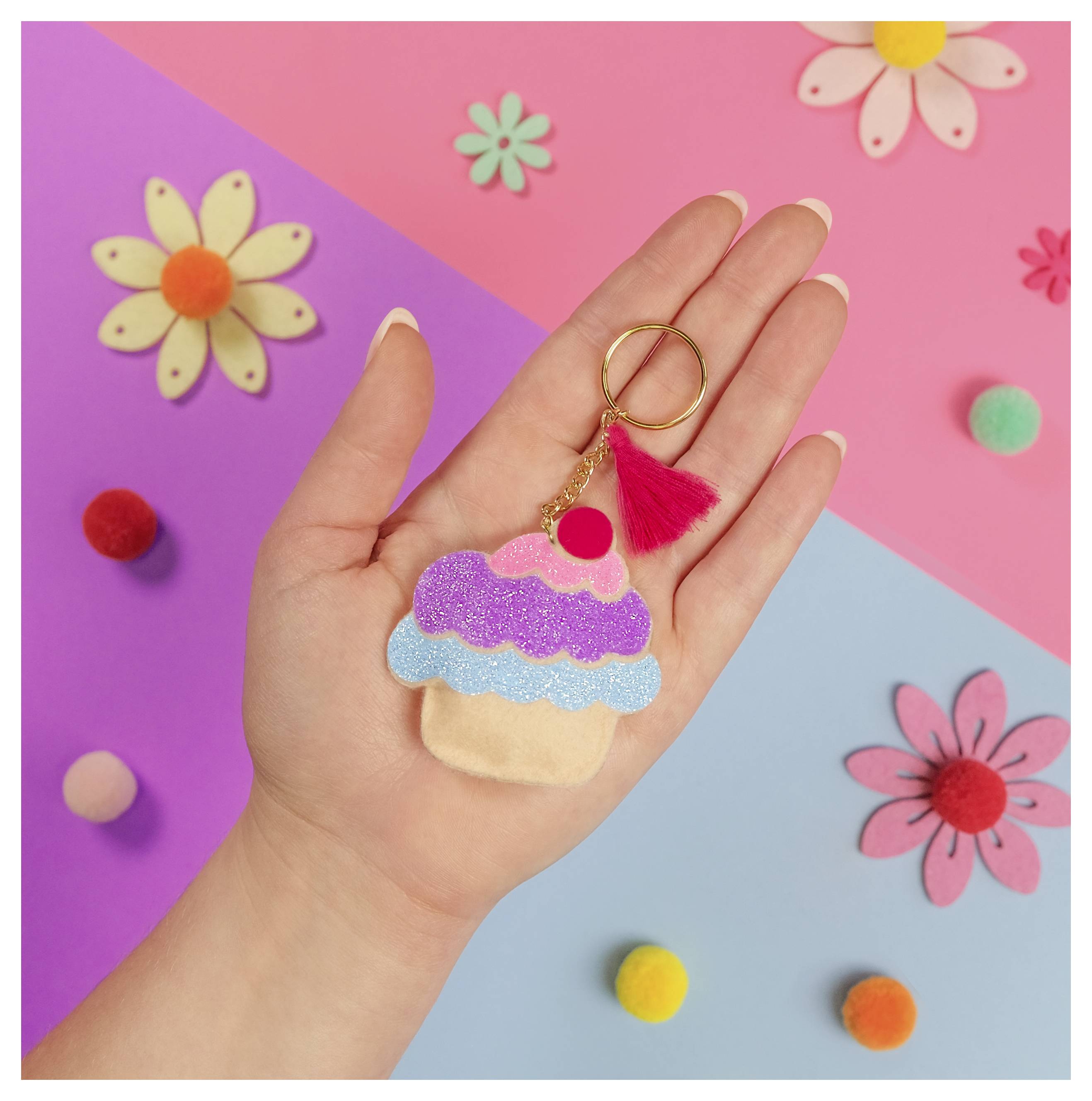 Ruka drží prívesok na kľúče v tvare cupcakea s ružlitým a modrým glitrom, umiestnený na farebnom pozadí s kvetmi a pomponmi.