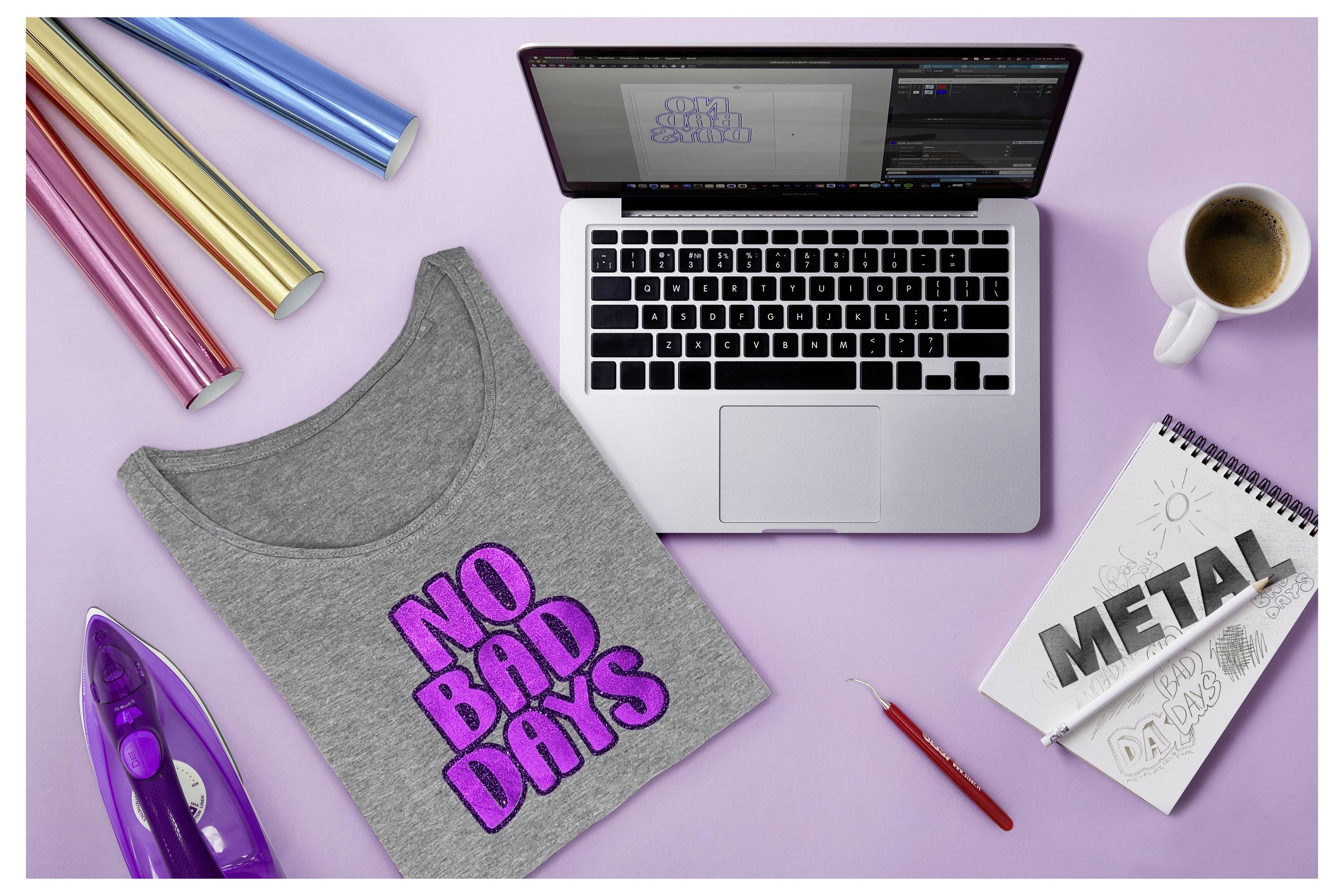 Laptop zobrazujúci softvér na dizajn, vedľa neho sivé tričko s nápisom „NO BAD DAYS" v ružovej farbe, žehlička, valce s vinylom, kávou a skicárom označeným „METAL".