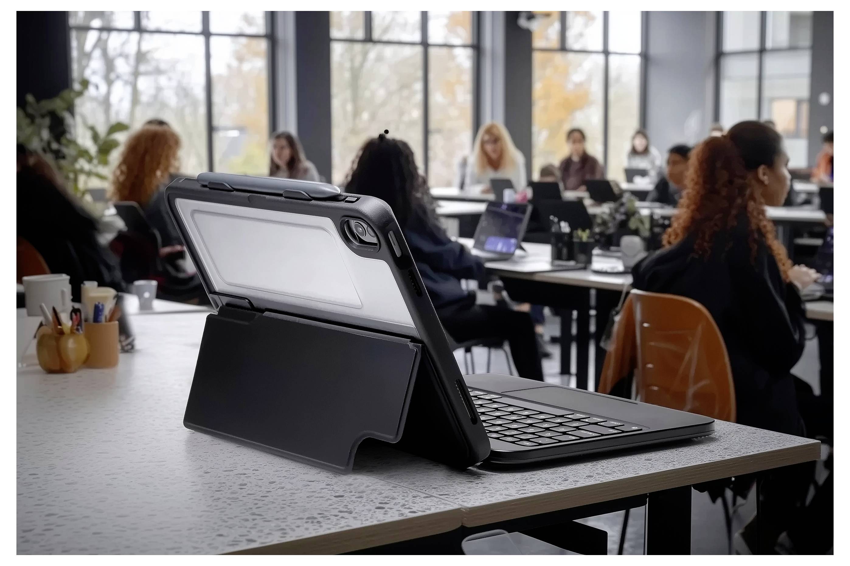Tablet s puzdrom na klávesnicu je na stole v popredí, v pozadí sú študenti sedení pri stoloch v prostredí učebne.