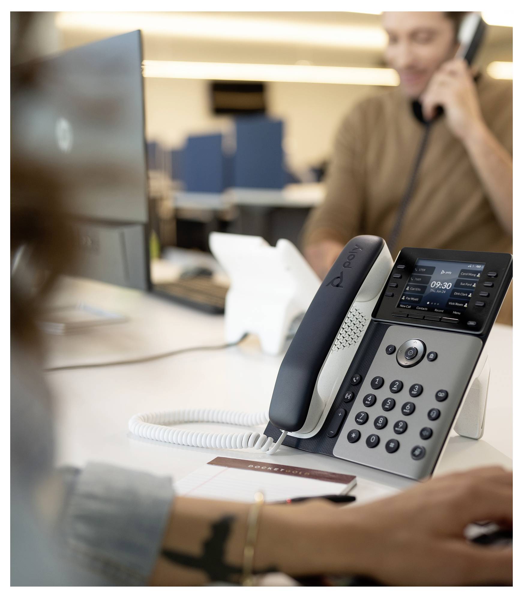 Telefón na pracovnom stole v popredí kancelárie s rozmazaným zamestnancom na hovoroch a počítačovým monitorom v pozadí.
