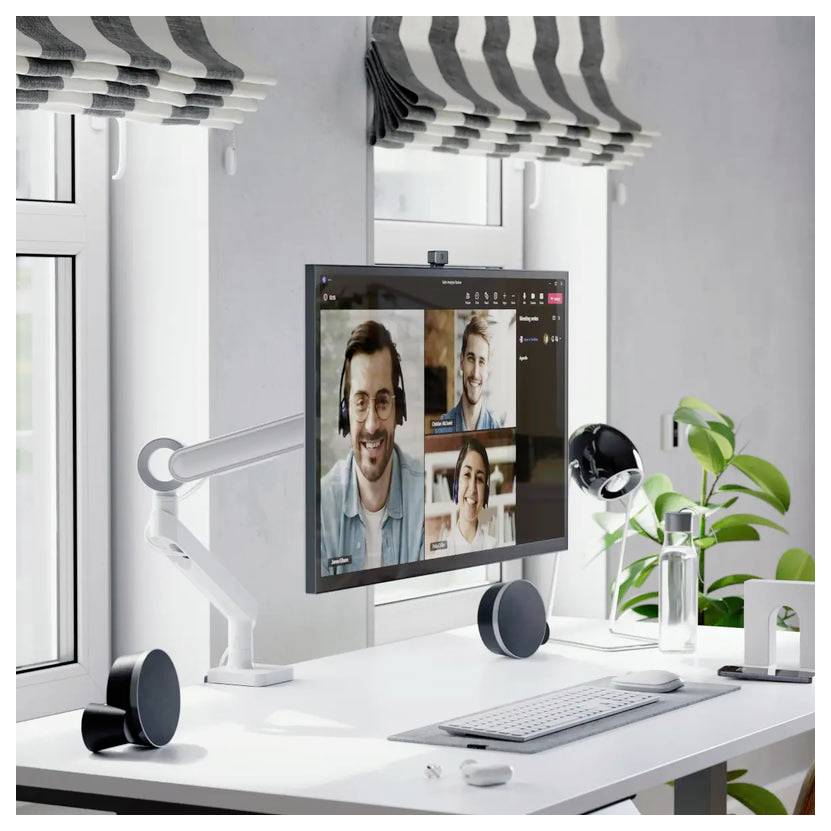 Kancelársky stôl na domáci kanceláriu s monitorom zobrazujúcim videohovor troch osôb, klávesnica, sluchátka, stolná lampa a kvetináč pri okne.