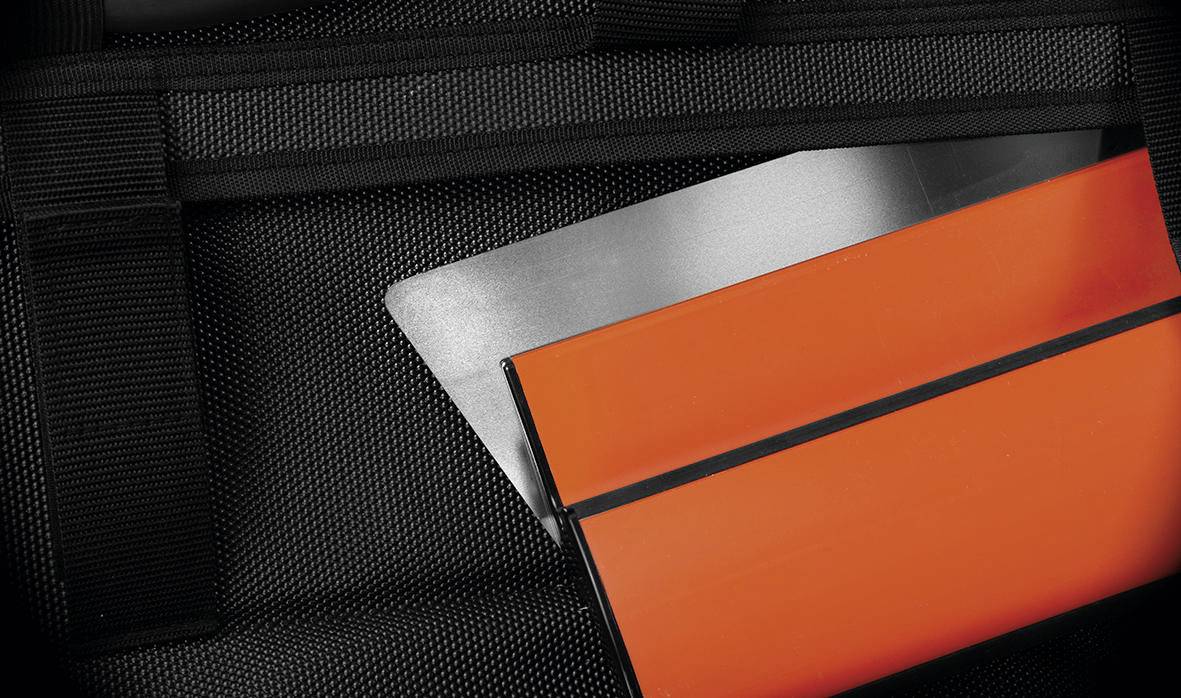 Detailný záber na oranžový a strieborný notebook čiastočne vložený do čierneho štruktúrovaného batožu, čo naznačuje cestovanie alebo dojčovanie.