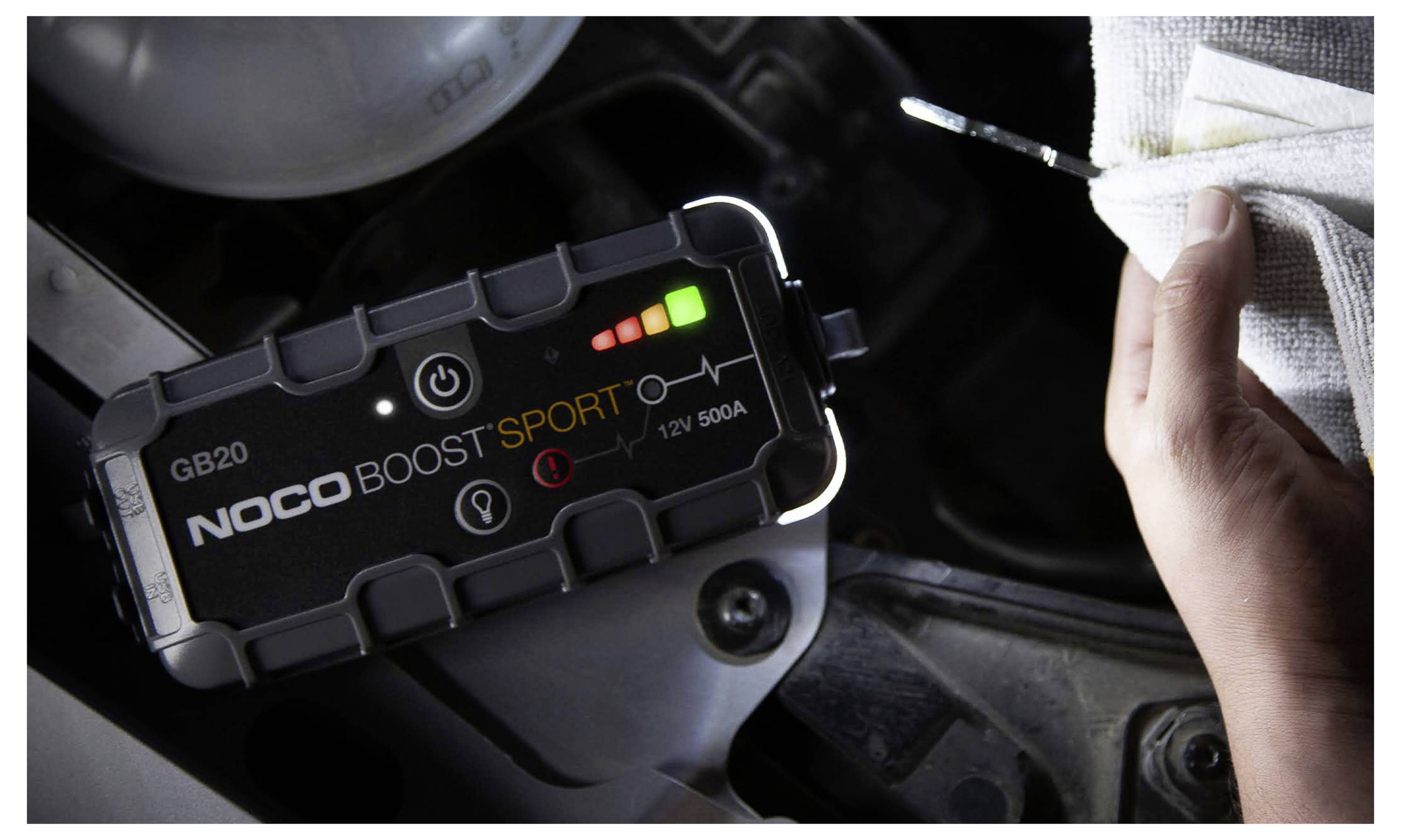 Prenosný štartér NOCO Boost Sport pripojený v motorovom priestore auta, s rukou držiavajúcou štartovacie kábly alebo vodiče v blízkosti.
