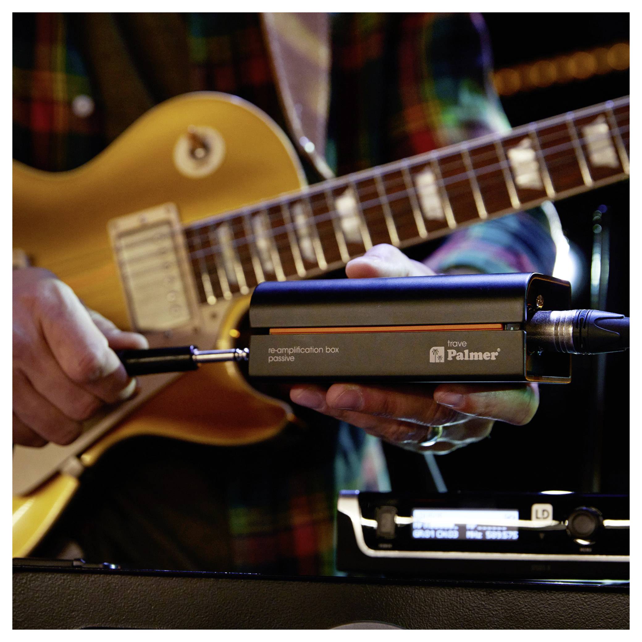 Osoba držiaca Palmer re-ampingový box pred elektrickou gitarou, zobrazujúca hudobné nahrávací alebo zosilňovacie zariadenie.