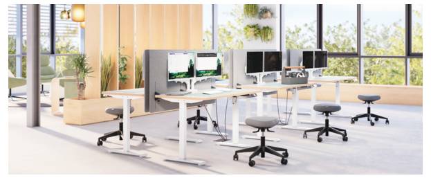 Moderná kancelária s otvoreným pôdorysom so stojacimi stolmi, viacerými počítačovými monitormi a otočnými stolčekmi pri veľkých oknách s rastlinami a teplým osvetlením.