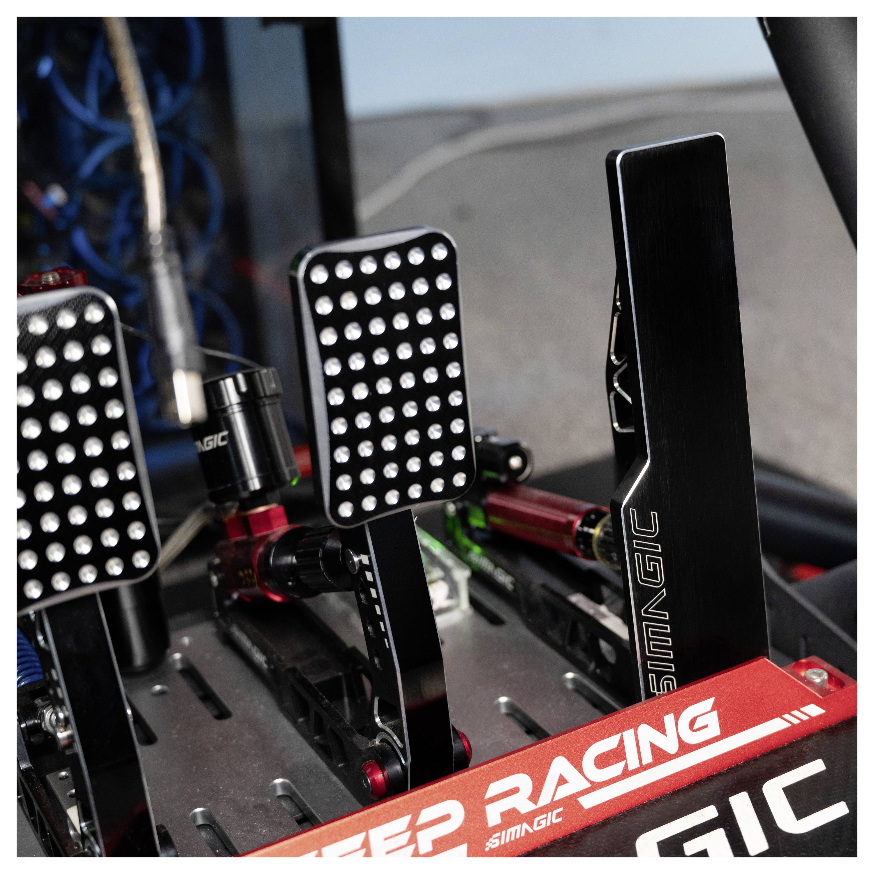 Zblízka zobrazená sada pedálov závodného simulátora s kovovými podložkami pre nohy, hydraulicky vyzerajúcimi valcami a červenou podpernou pákou s nápisom "RACING".