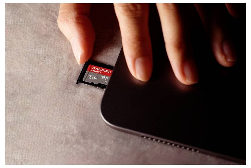 Ruka vkladajúca kartu SanDisk microSD vedľa notebooku, zobrazujúca rozšírenie úložného priestoru pamäťovej karty.