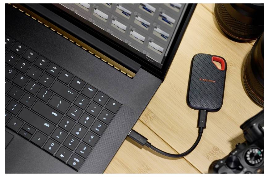 Pohľad zhora na notebook pripojený kábľom k prenosnej externej SSD jednotke SanDisk na drevenom stole s fotografickým vybavením v blízkosti.