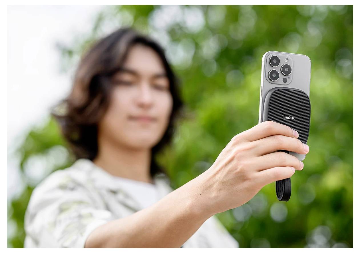 Osoba si robí selfie vonku a drží smartfón s čiernym magnetickým držiakom pred rozmazaným zeleným pozadím.