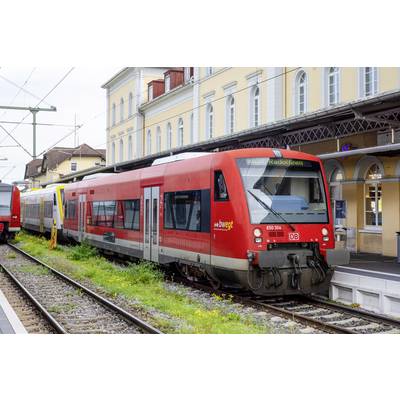 Červený regionálny vlak na staničnej plattforme s vzdušnými vedeniami a historickými budovami v pozadí.