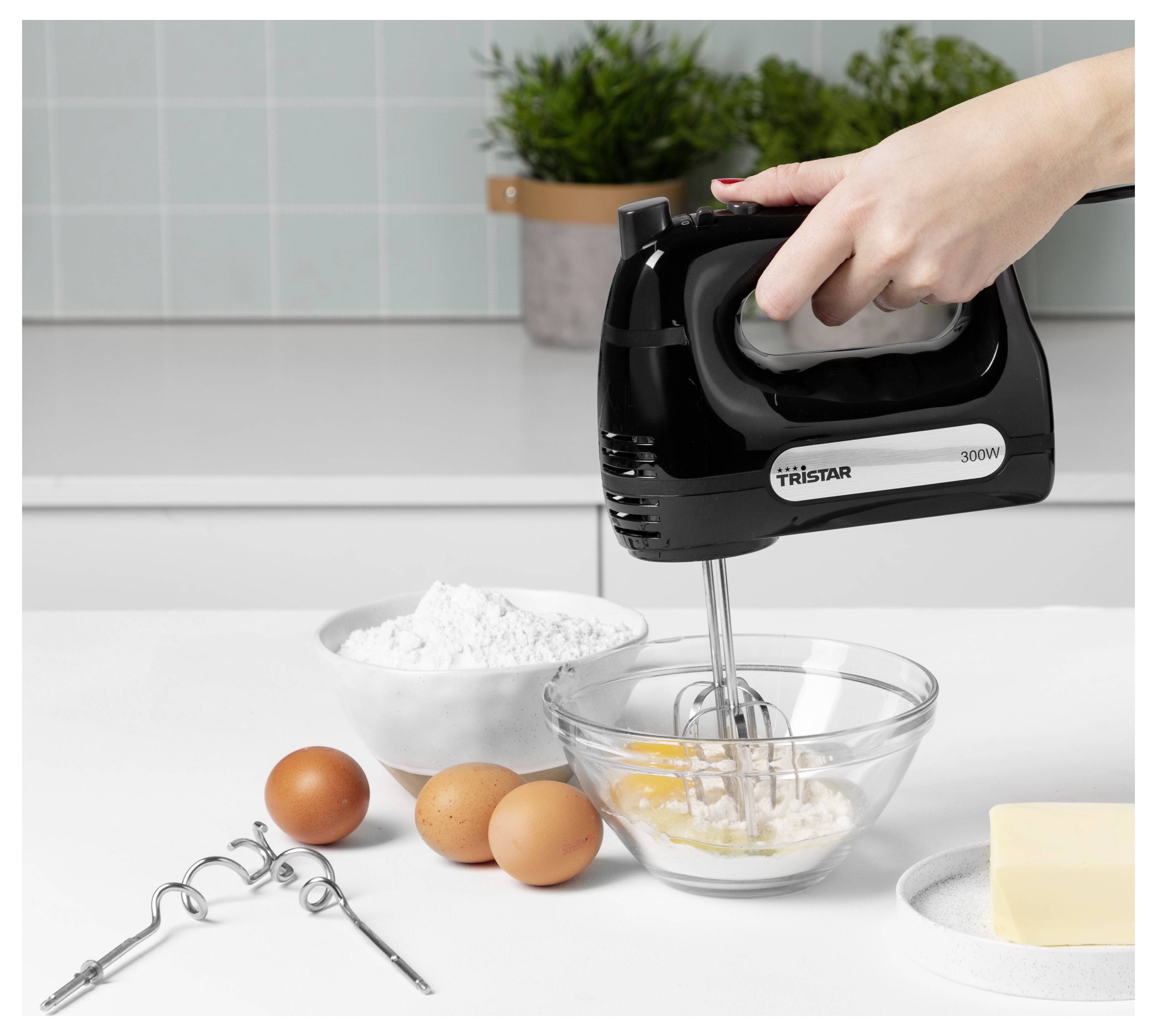Ručný mixer miešajúci ingrediencie v sklenenej miske na kuchynskom pulte, v blízkosti vajec, múky, metličiek a masla.