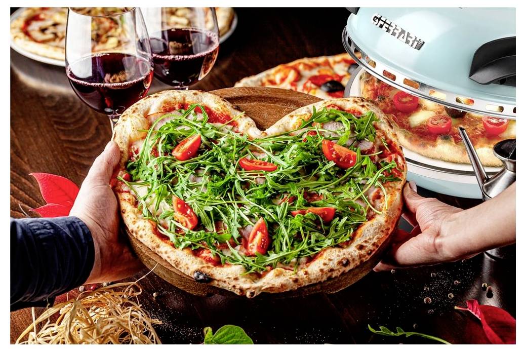 Ruky držiace pizzu v tvare srdca z rukoly a paradajok na drevenom prkne, s pohármi vína a pecou na pizzu v pozadí.