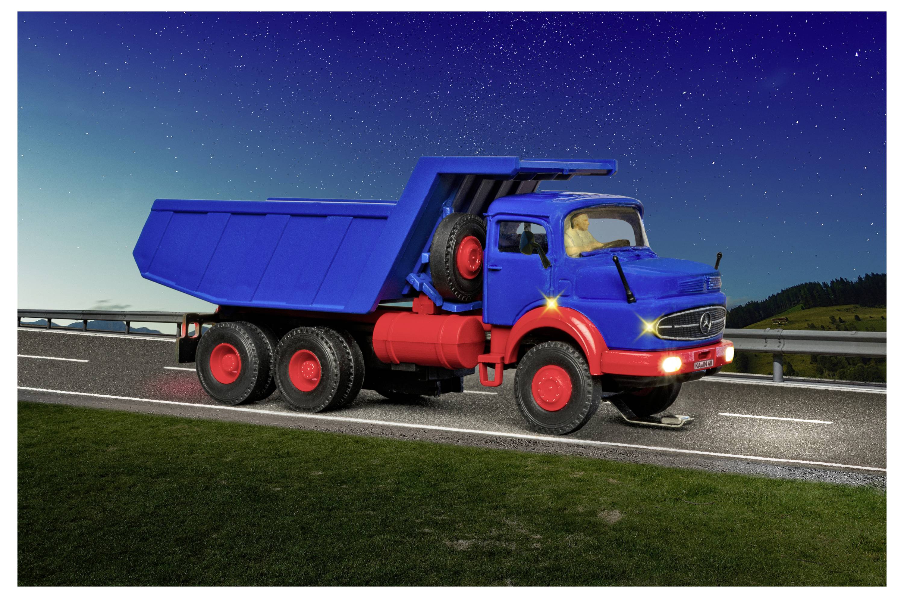 Modrý skladací nákladný automobil zaparkovaný na ceste pri súmraku s zapnutými predními svetlami a hviezdnatou oblohou v pozadí.