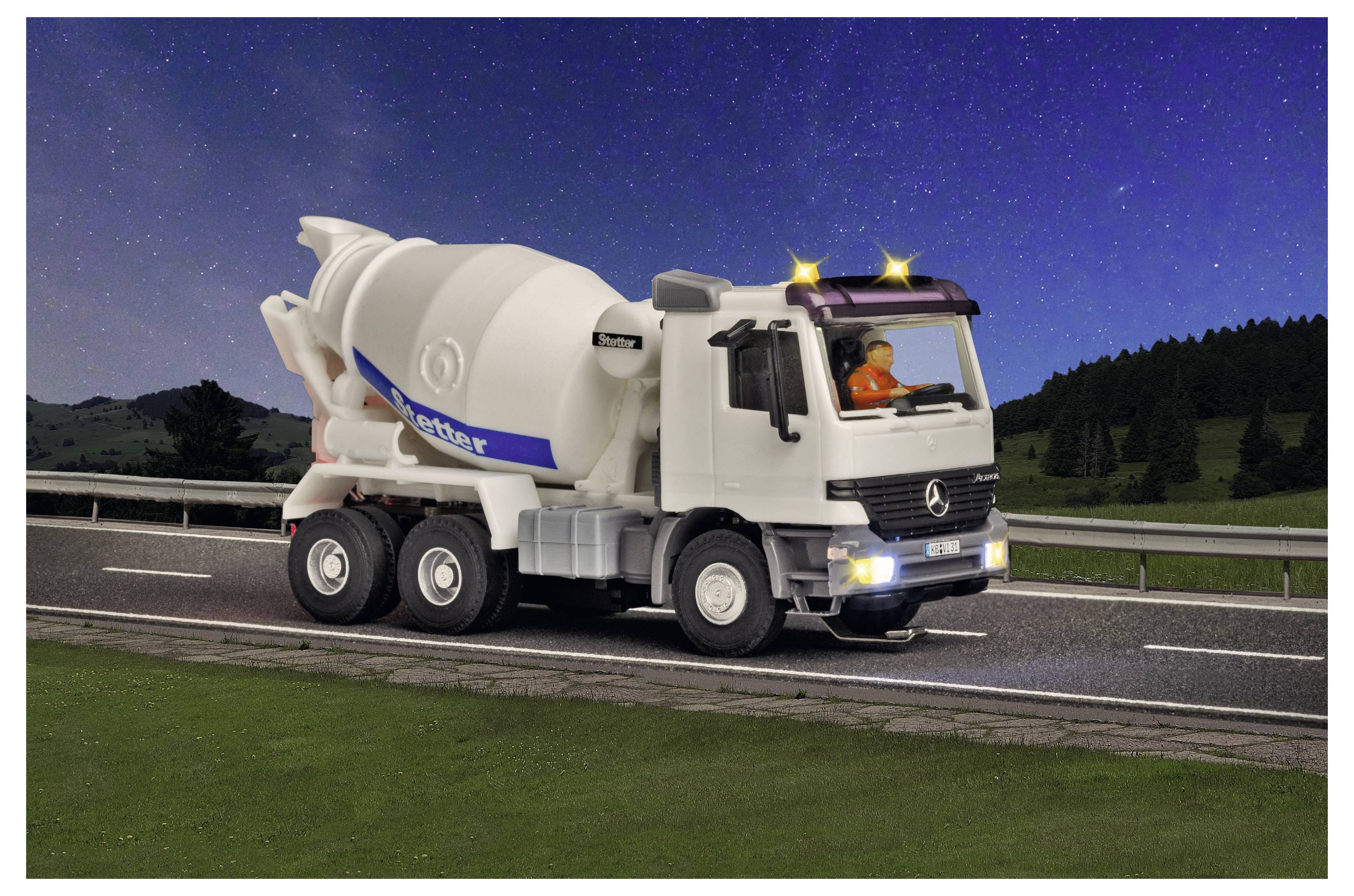 Biely cement mixer truck jazdia na diaľnici v noci so svietidlami a strechovými svetlami zapnutými, pred tmavou modrou oblohou a kopcami pozdĺž cesty.