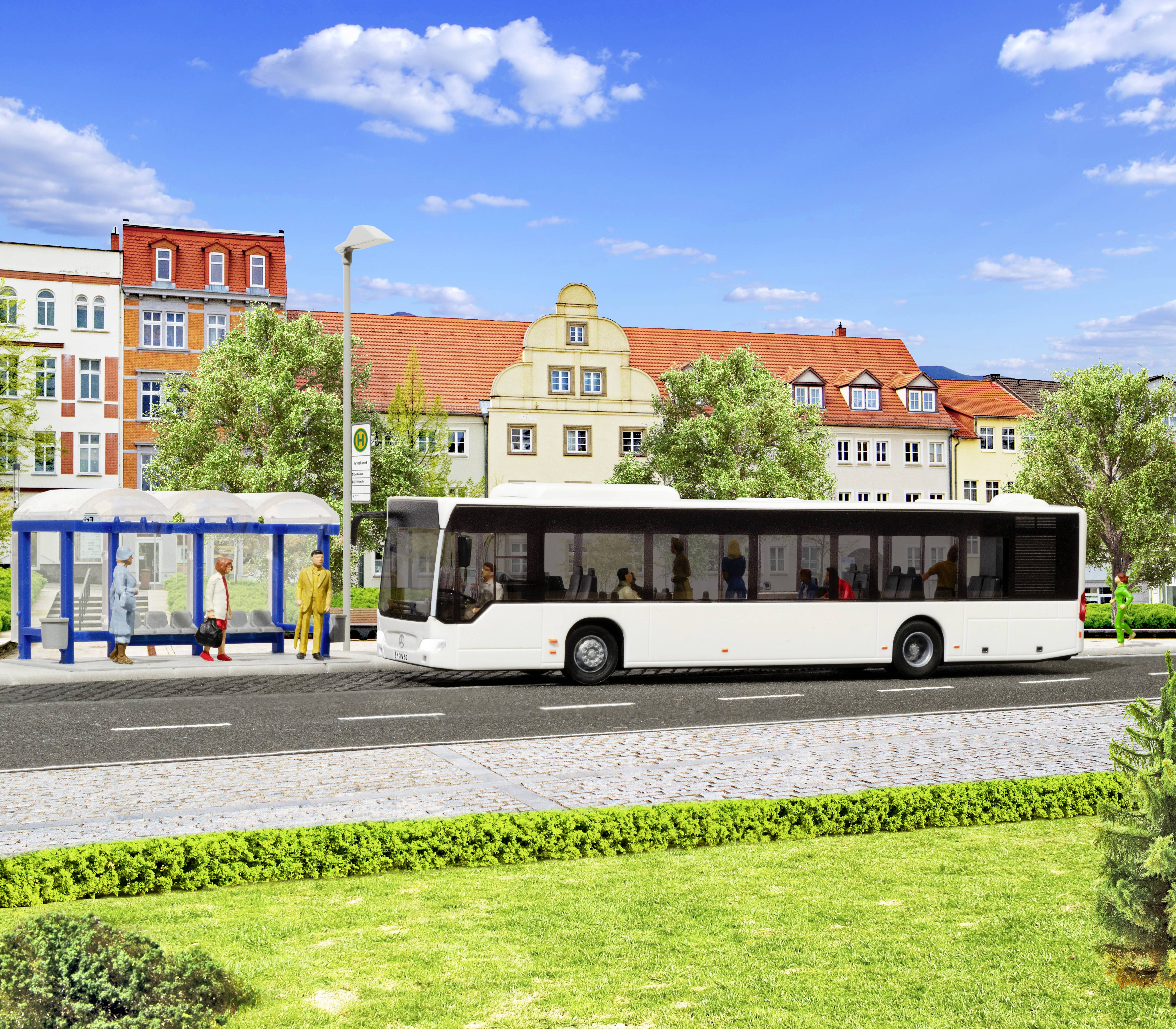 Biely mestský autobus zastavil vedľa krytej autobusovej čakárne na ulici so zafarbeными budovami a stromami v slnečný deň.