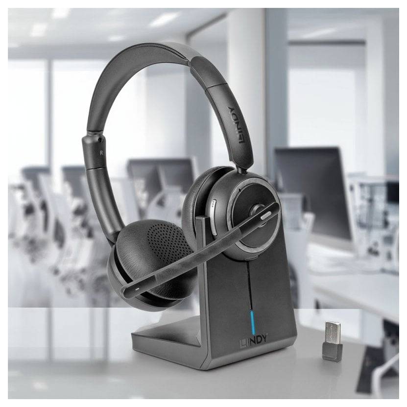 Čierny headset cez ucho s výbavou dynamického mikrofónu na stojacej nabíjačke, zobrazený na pracovnom stole v rozmazanom kancelárskom prostredí s USB donglom vedľa neho.