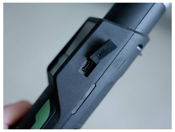 Zblízka čierneho ručného zariadenia s portom micro-USB na nabíjanie a neďalekým spínačom, so zeleným akcentom na rukoväti.