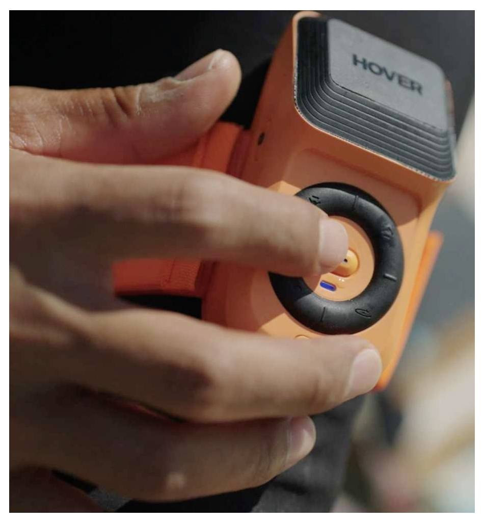 Zblízka ruka tlačiaca na ovládací gombík na oranžovom zariadení HOVER.
