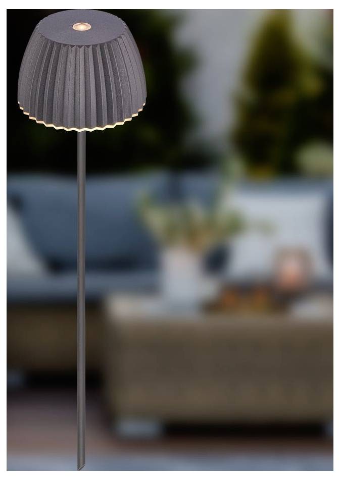 Sivá záhybová stojaca lampa s teplým svetlom, stojaca pred rozmazanou obývacou izbou s pohovkou a stolom.