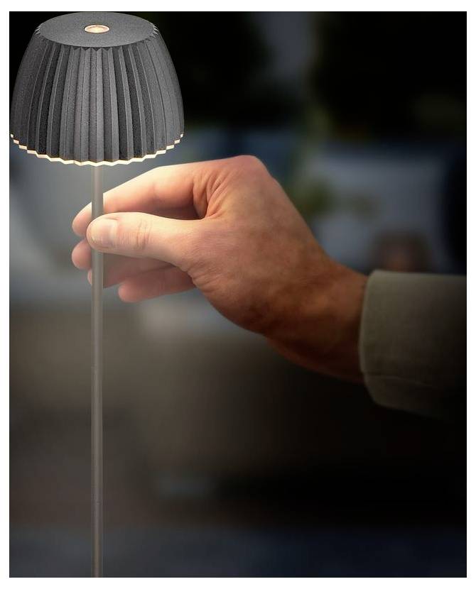 Ruka upravujúca malú sivú lampu s vrásovaným tieňom, čo naznačuje stolnú alebo modelovú svetelnú armatúru.