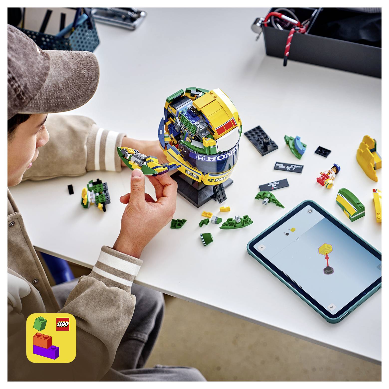 Dieťa staviace prilbu z LEGO modelu pri stole podľa pokynov na tablete, s rozptýlenými kosťami a časťami v blízkosti.