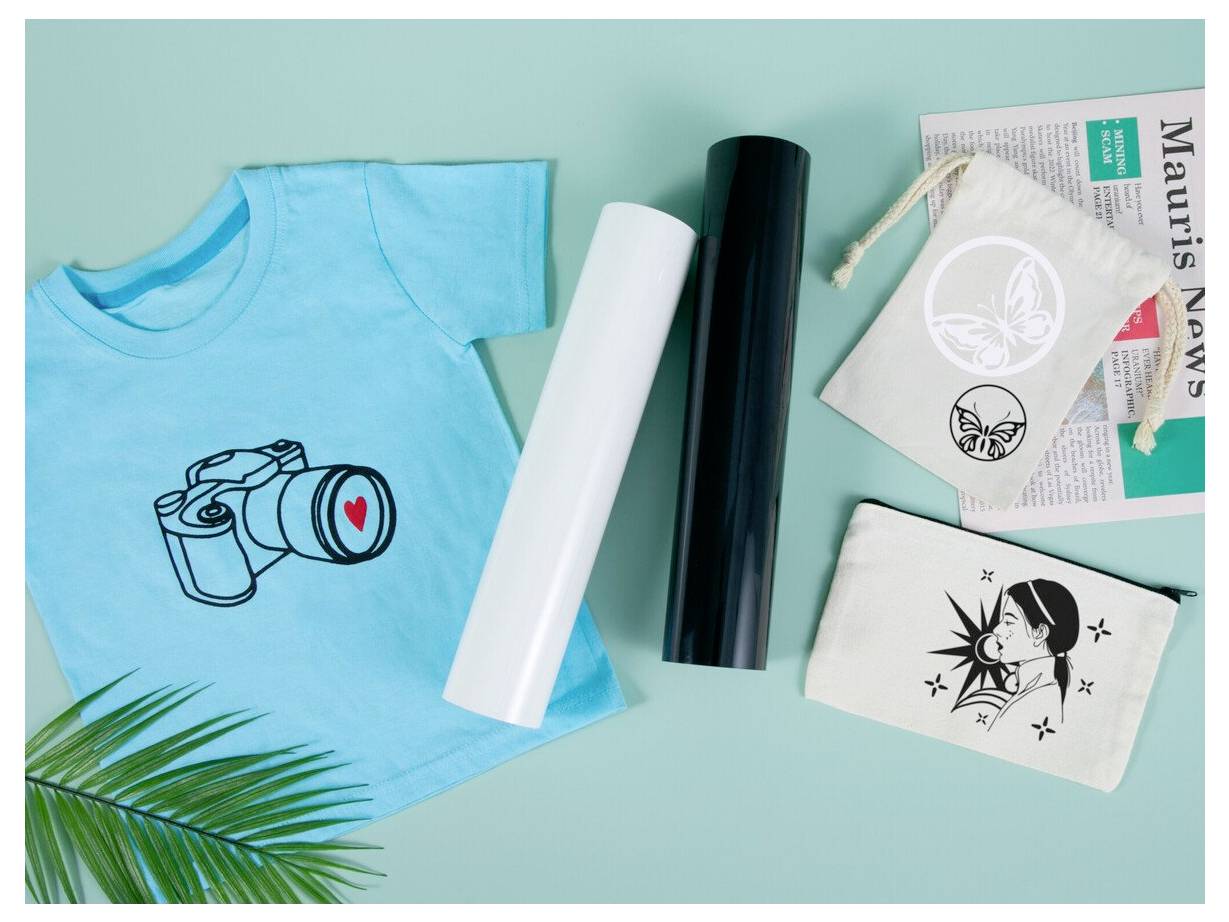 Flat-lay zobrazenie tyrkysového trička s grafickou potlačou fotoaparátu, dvoch vinylových transferových pásk, vytlačených nálepiek a malej vrecúška na mätovom pozadí.