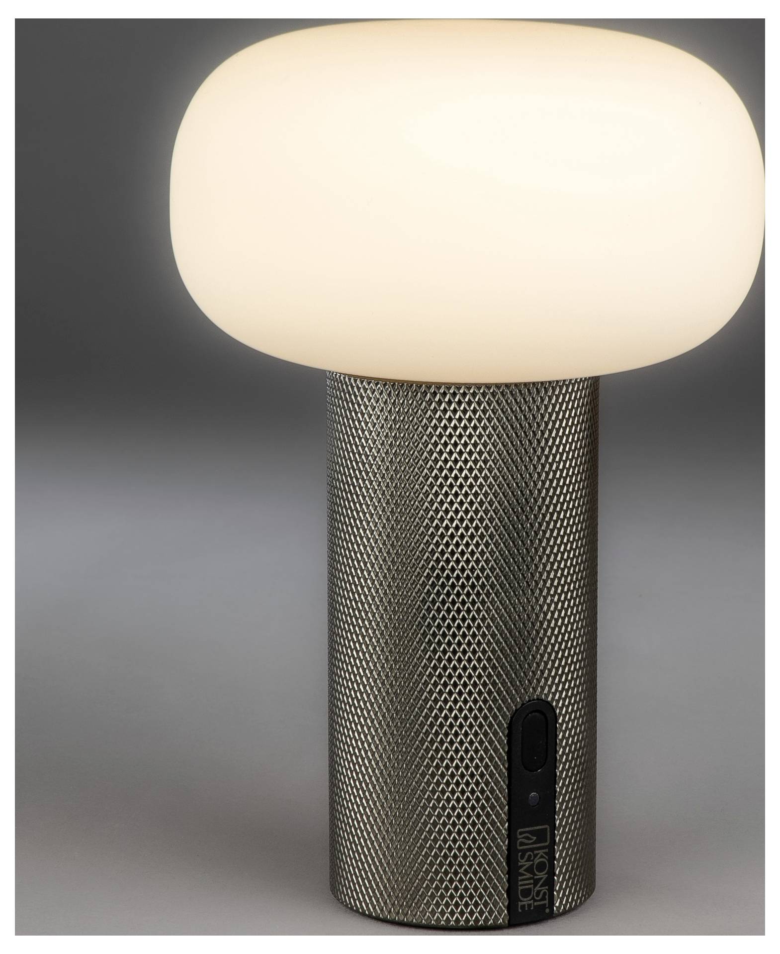 Moderná bezdrôtová stolová lampa so svietiacim zaoblenú tienidlom a štruktúrovanou kovovou základňou, zobrazená na jednoduchom sivom pozadí.