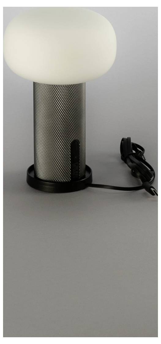 Moderná stolová lampa s bielym zaoblenám tienidlom, perforovanou kovovou valcovitou podstavou a pripojeným elektrickým kábľom na sivom pozadí.