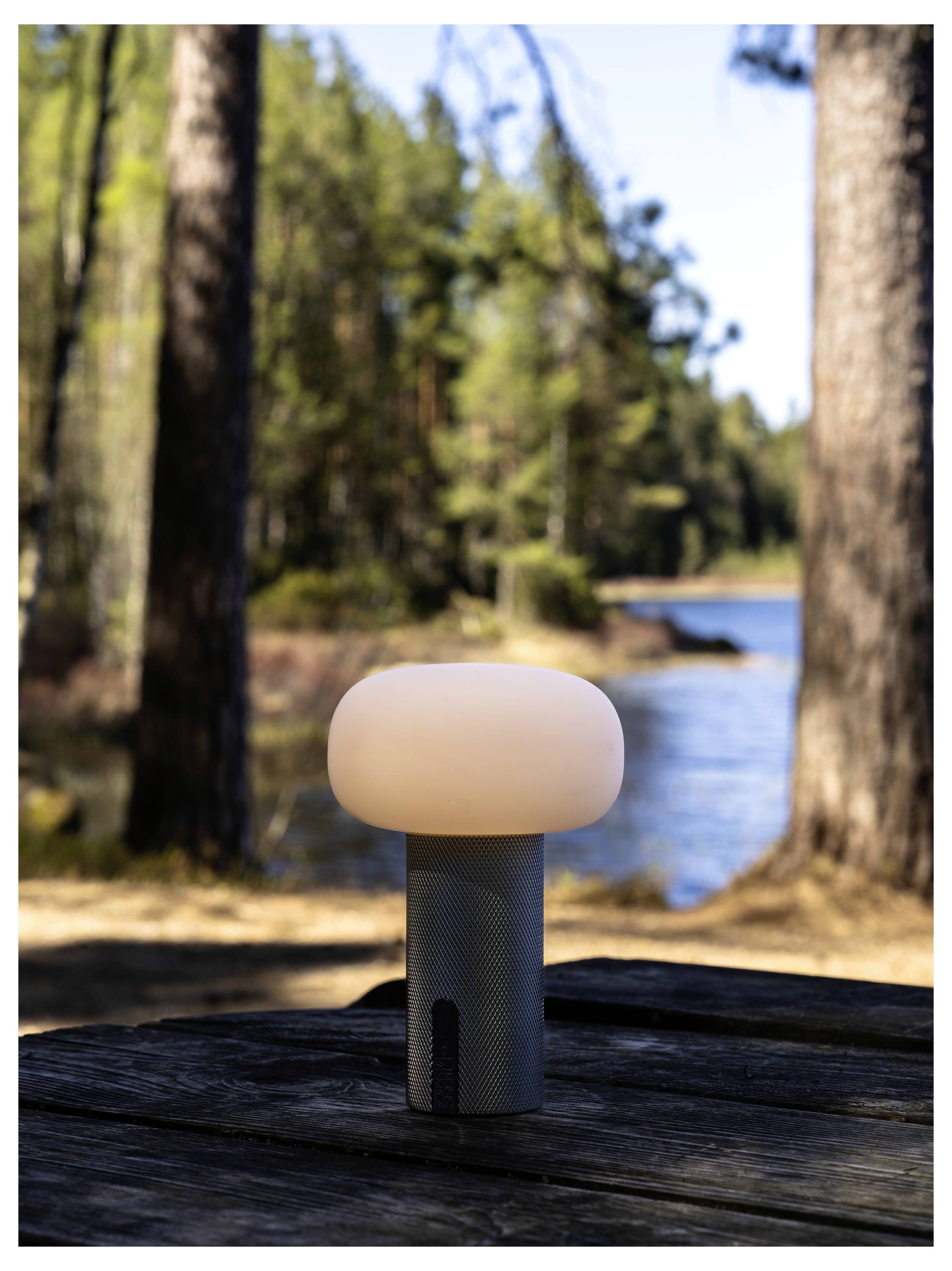 Malá valcovitá lampa so zaoblenským bielym vrchom stojí na drevenom stole vedľa rieky v lesnatej oblasti.