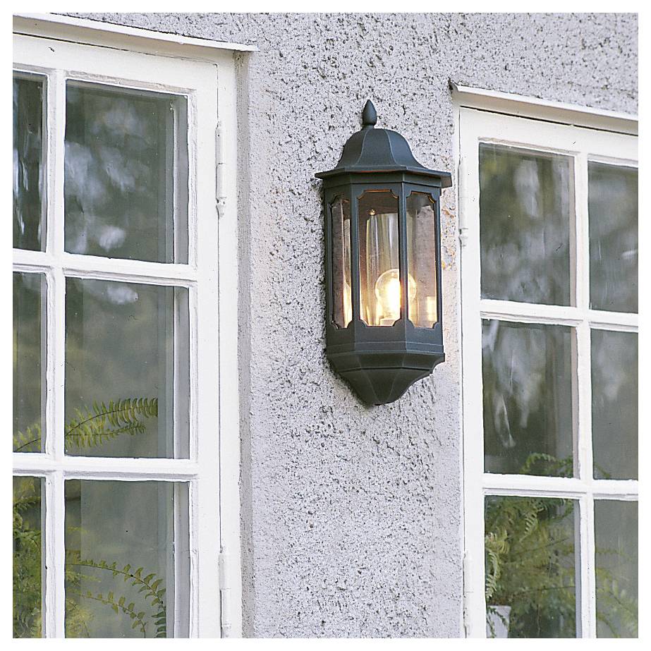 Vonkajšia nástenná lampa rozsvietená medzi dvoma oknami s bielym rámom na omietanej budove.