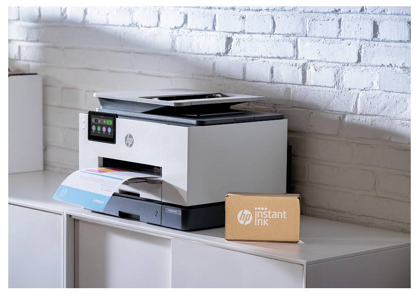 Tlačiareň HP na skrinke s farebným výtlačkom a krabičkou „HP Instant Ink" vedľa nej na pozadí bielej tehlickej steny.