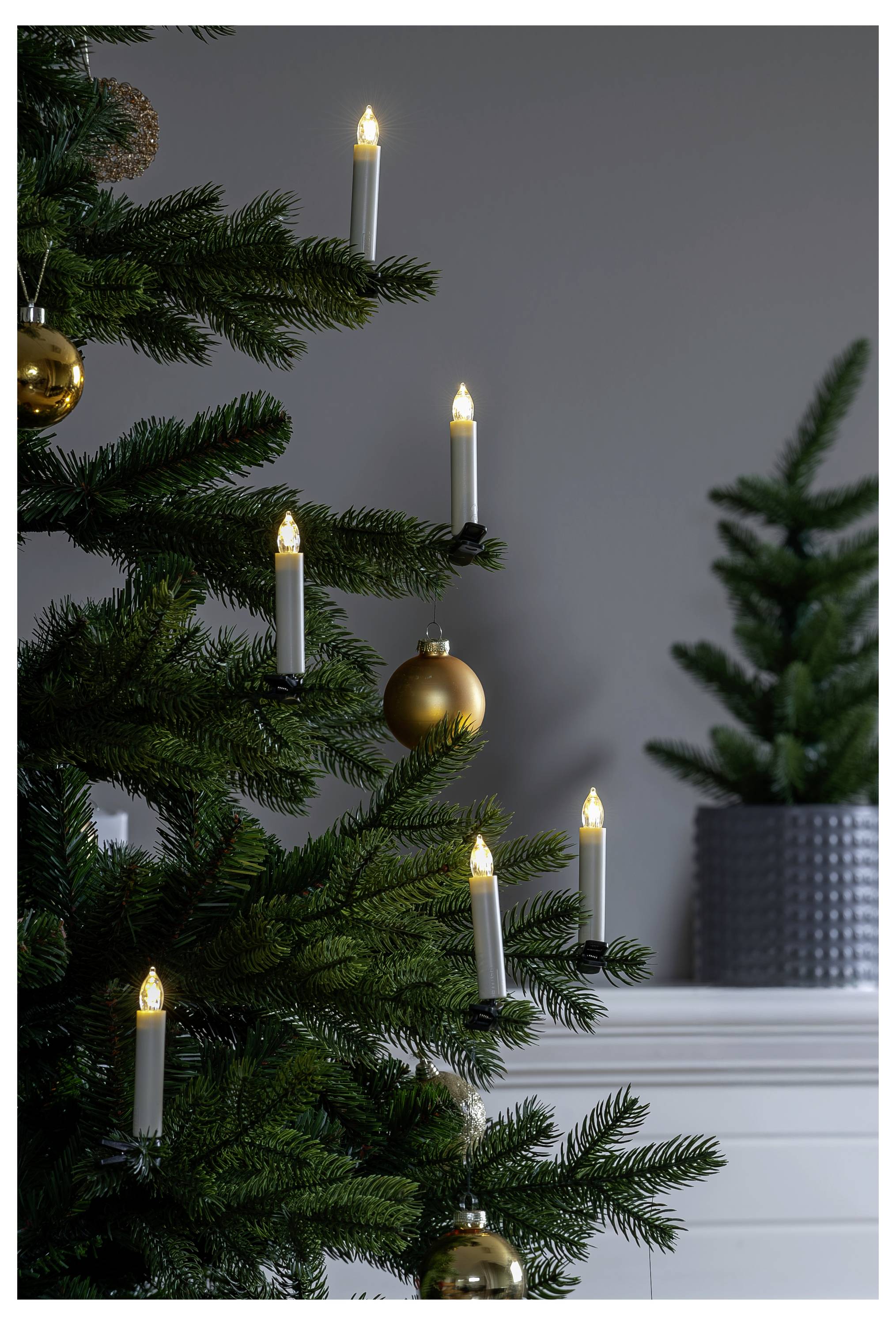 Zdobia Christmas stromček s rozžiarenými bielymi sviečkami a zlatými ozdobami vedľa bieleho krbu pri sivej stene.