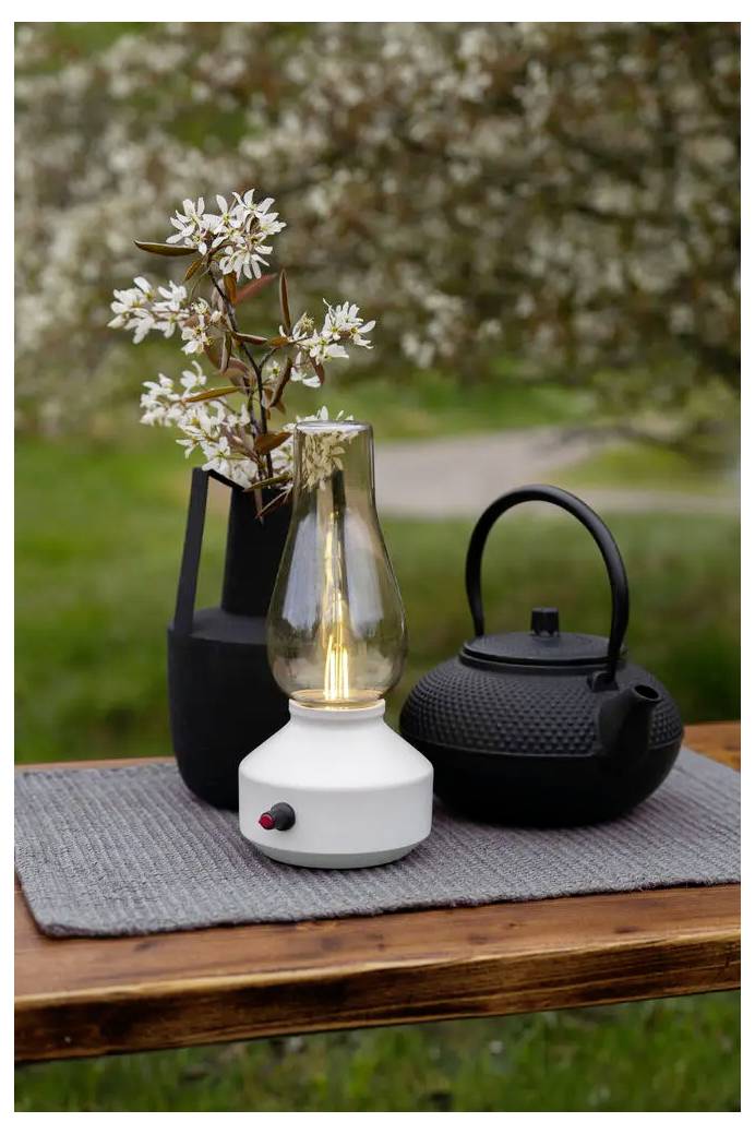 Čierna kanvica, zapnutá biela stolová lampa a váza s bielymi kvetmi usporiadané na drevenom stole vonku.