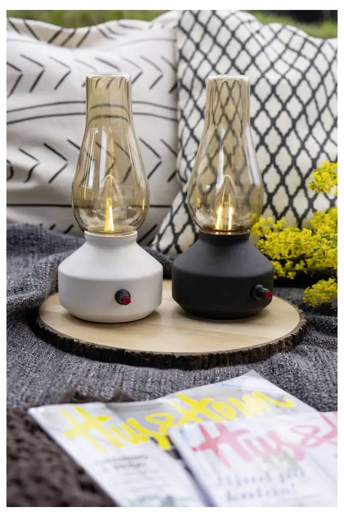 Dva malé prenosné lampáše na dreve s rozžiarenými žiarovkami, v bielych a čiernych podstavcoch, stylizované na pohodlnej deke so vzorovými vankúšmi a kvetmi.