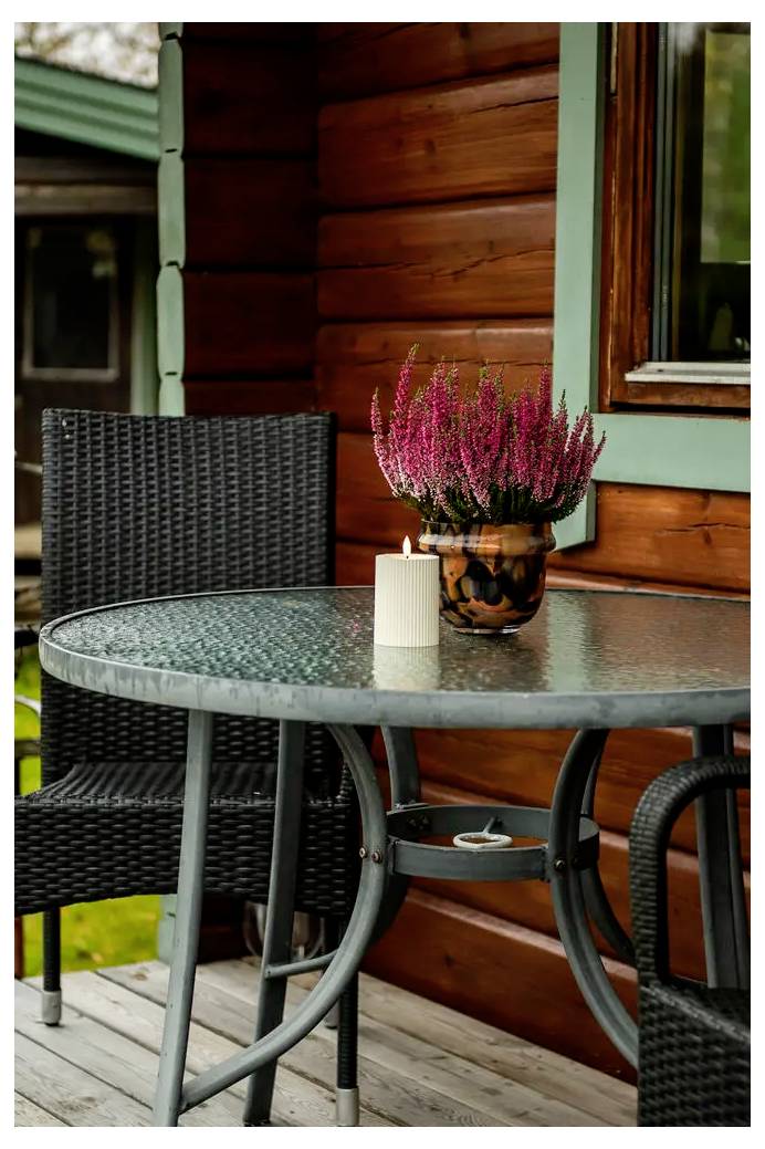 Vonkajší patiovací stôl so sviečkou a kvetináčom ružových kvetov vedľa prútených stoličiek pri drevnej stene chaty