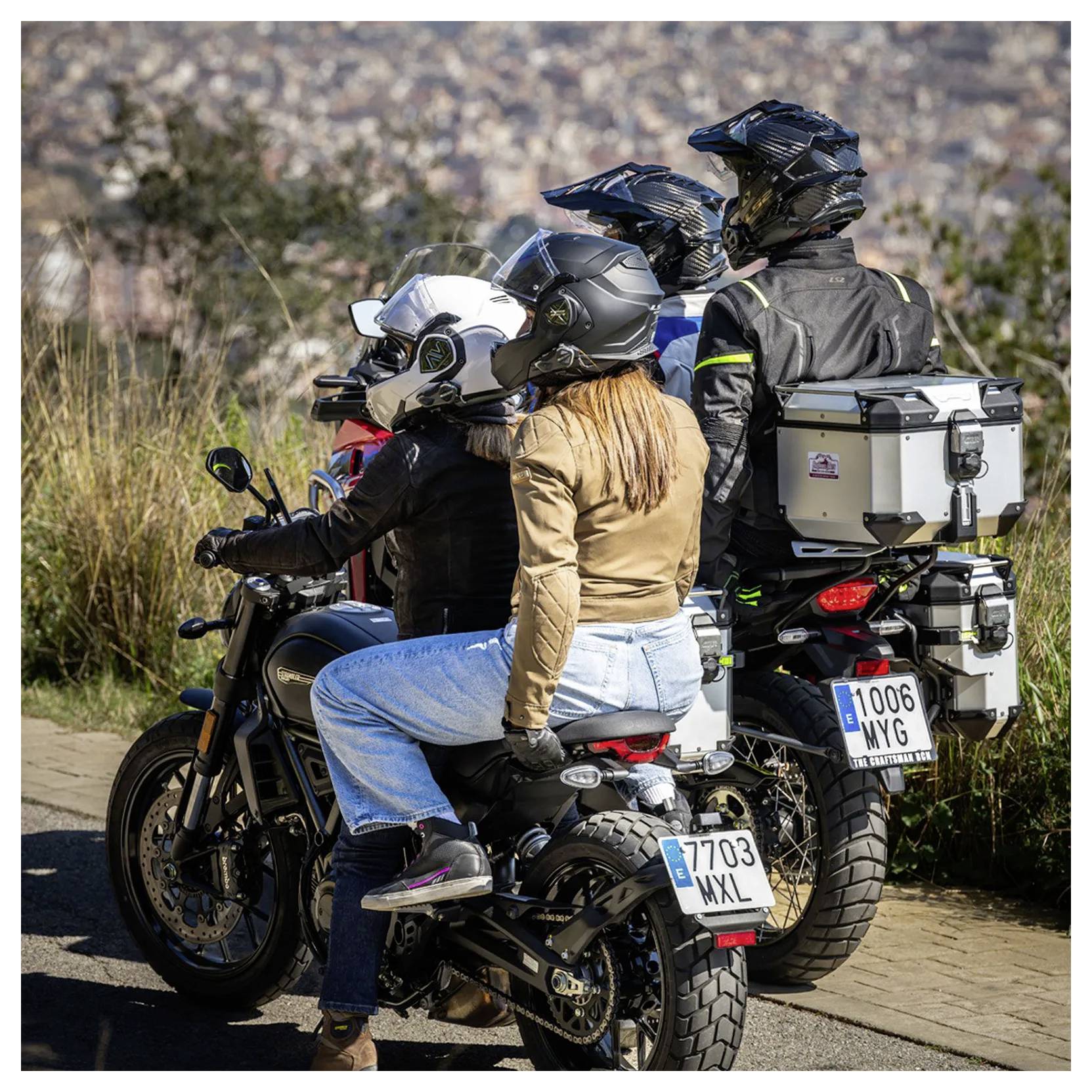Traja motorkári v prilbách čakajú na adventúrnych motorkách pri okraji cesty, pričom na zadnom motocykli sú pripevnené putovné kufre.