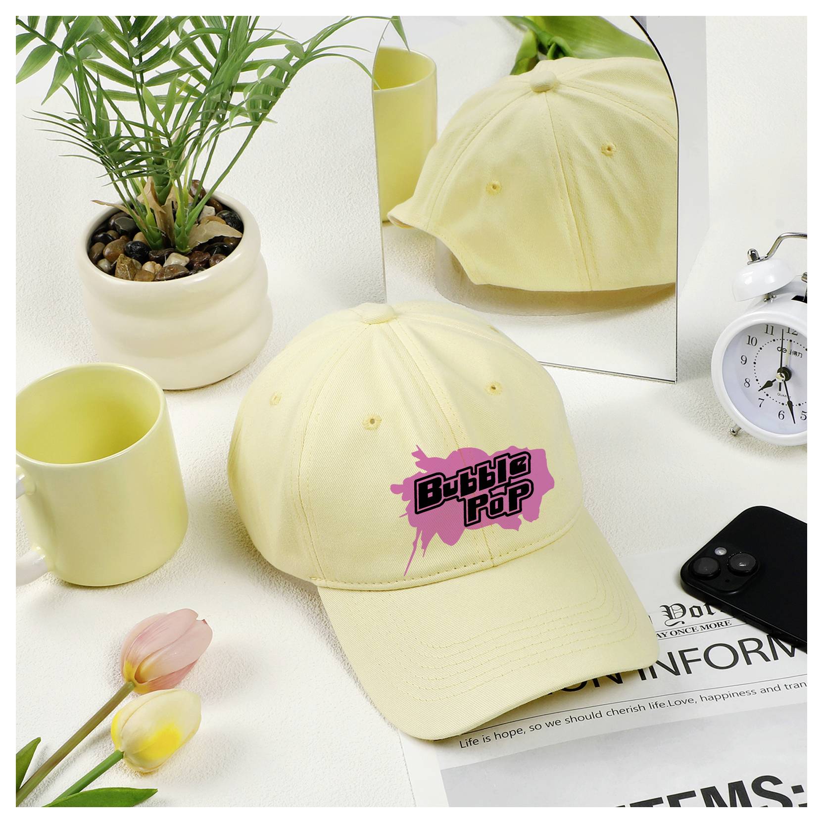 Svetložltá baseballová čiapka s logom „Bubble Pop", zobrazená na bielej stolnej doske vedľa rastliny, sviečok, kvetov, hodín a odrazu v zrkadle.