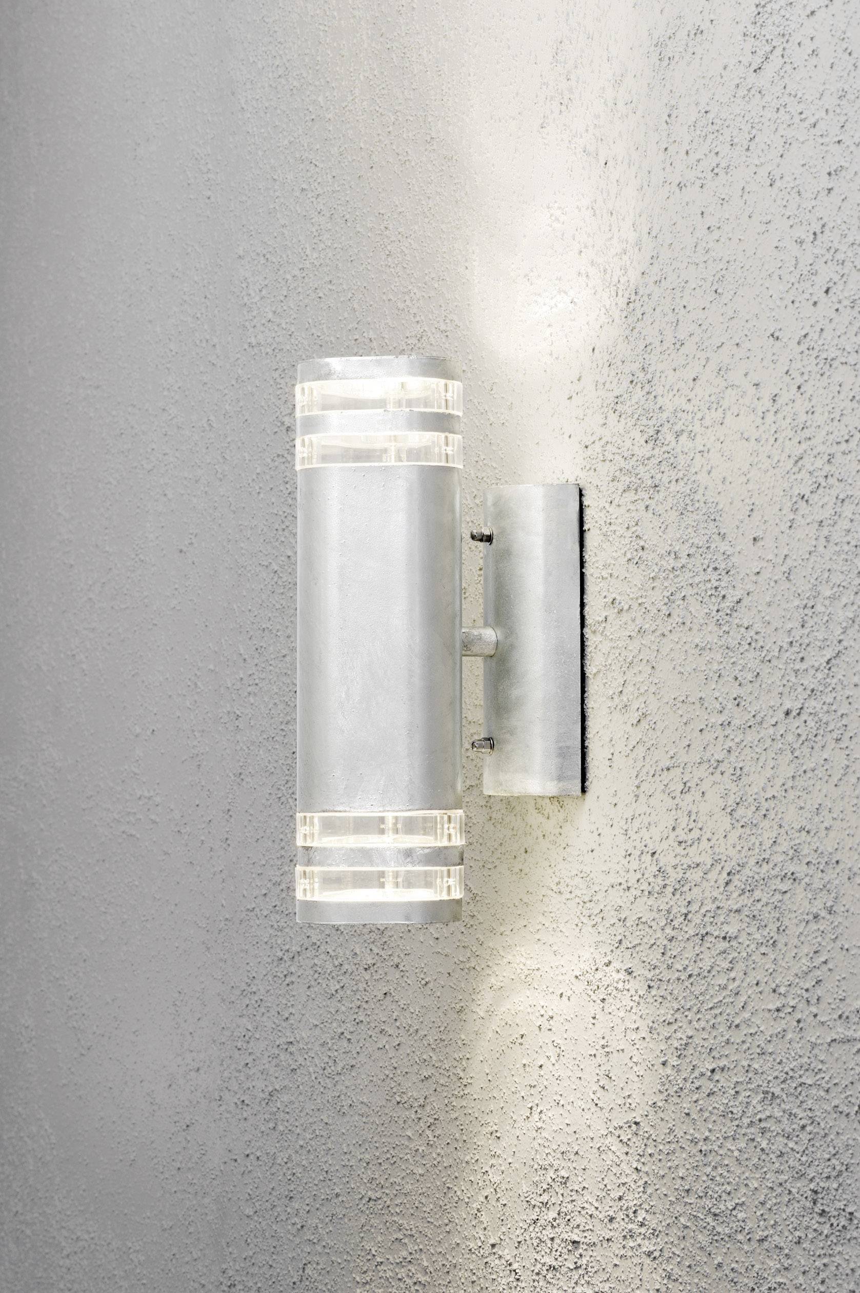 Strieborná valcovitá vonkajšia lampa pripevnená na sivej stene, ktorá svieti nahor a nadol a jemne osvecuje stenu.