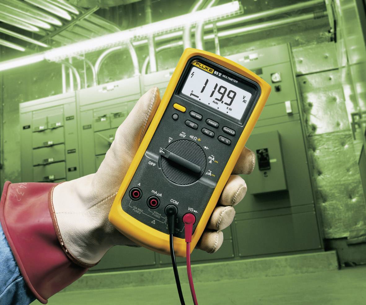 Jedna ruka drží žlté multimeter s digitálnym displejom, ktorý zobrazuje '119,9 V'. V pozadí sú viditeľné elektrické rozvádzače.