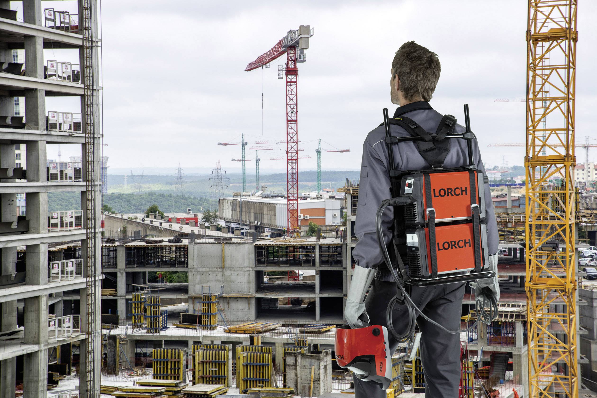 Robotnícky pracovník v exoskelete sa pozerá na stavenisko s viacerými žeriavmi a budovanými objektmi.