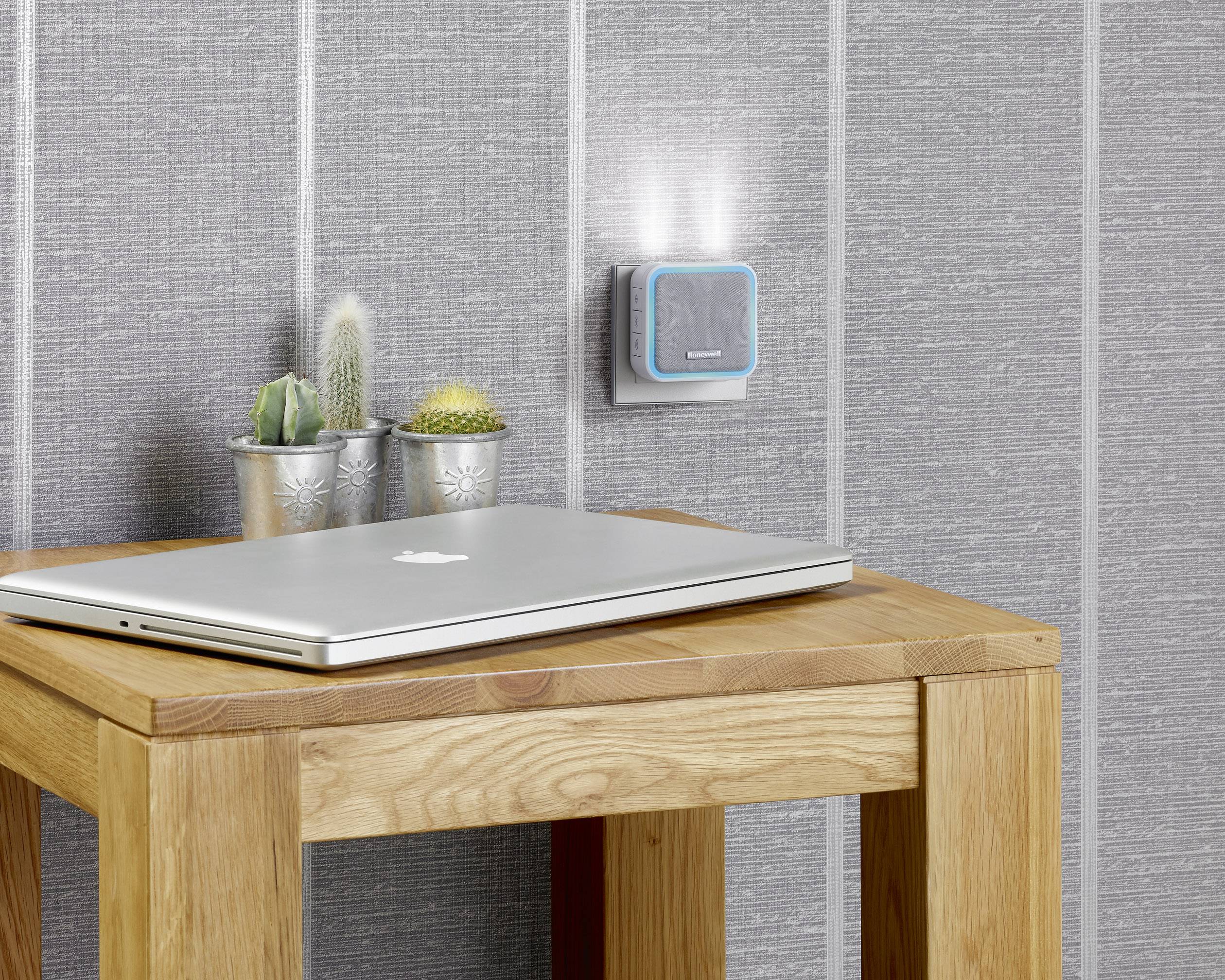 Laptop leží na drevenom stole vedľa dvoch malých kaktusov. Na sivej stene je umiestnený osvetlený spínač.