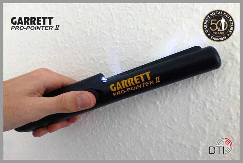 Jedna ruka drží čierny kovový detektor s názvom 'Garrett Pro-Pointer II', ktorý je používaný so svetlom pri bielej stene.