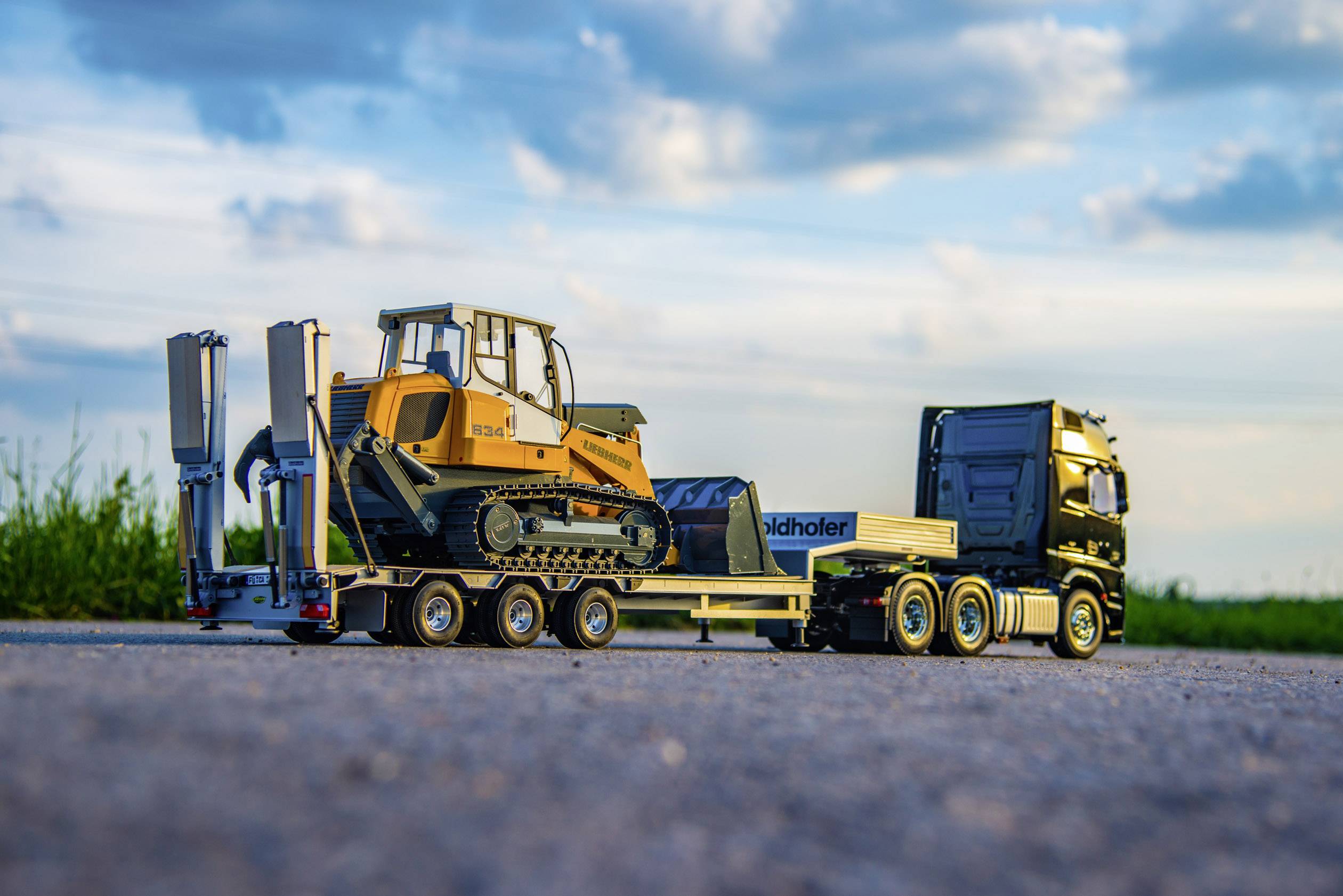 Žltý nákladný automobil s prívesom prepravuje veľký bager po poľnej ceste počas západu slnka.