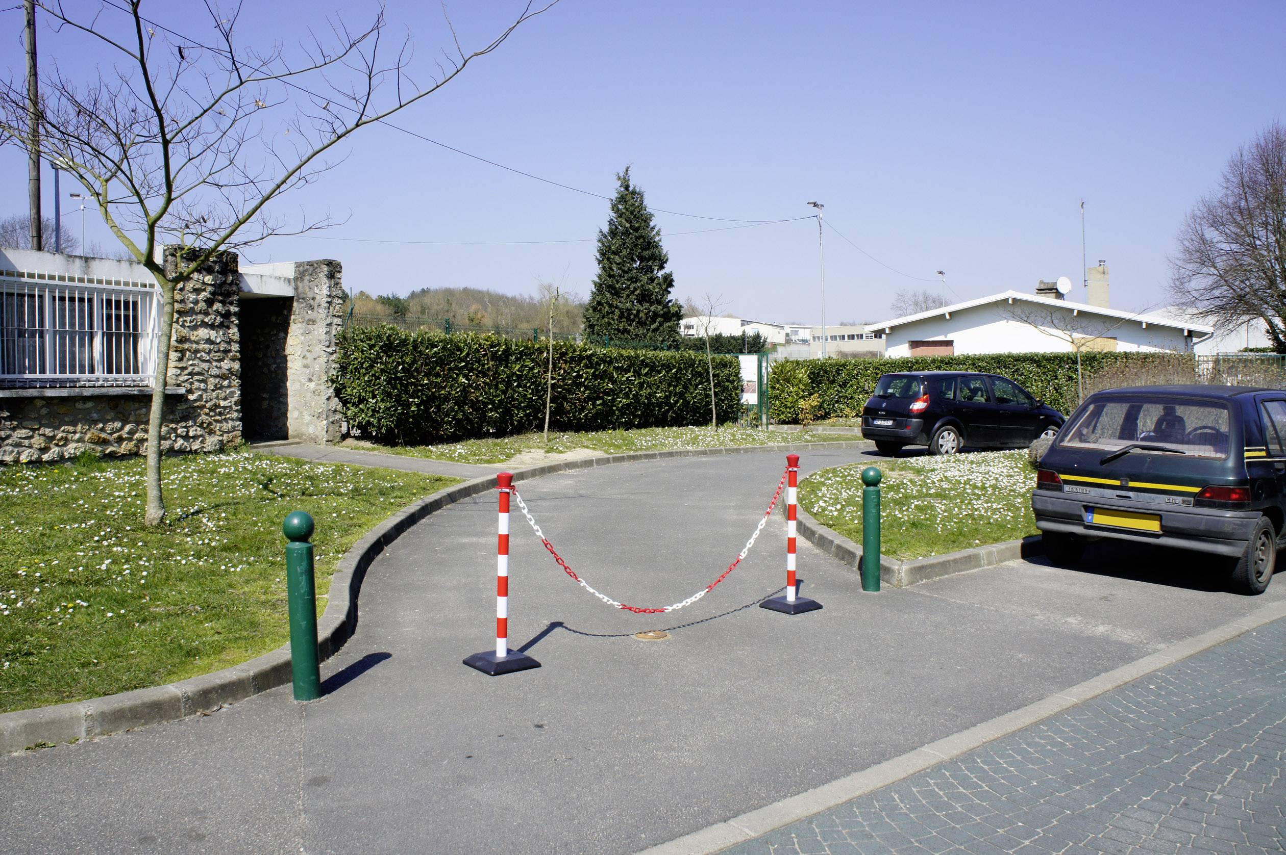Dva automobily sú zaparkované na malom parkovisku, ktoré je čiastočne blokované červeno-bielymi zábranami. V pozadí je strom a kríky.