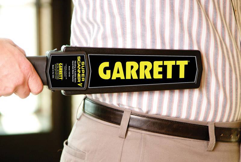 Osoba je kontrolovaná ručným kovovým detektorom značky Garrett po strane hornej časti tela.